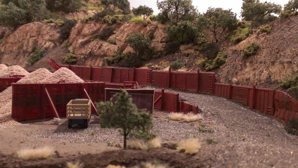 THIS SCENE DEPICTS A TYPICAL AGGREGATE LOAD OUT FACILITY FROM TRUCK TO RAIL. SPARE PARTS ERE USED TO CONSTRUCT RETAINING WALLS FOR THE GRAVEL, AND IN THE BACKGROUND YOU CAN SEE A ROCK OUTCROPPING AND TREES TYPICAL OF NORTHERN CALIFORNIA.